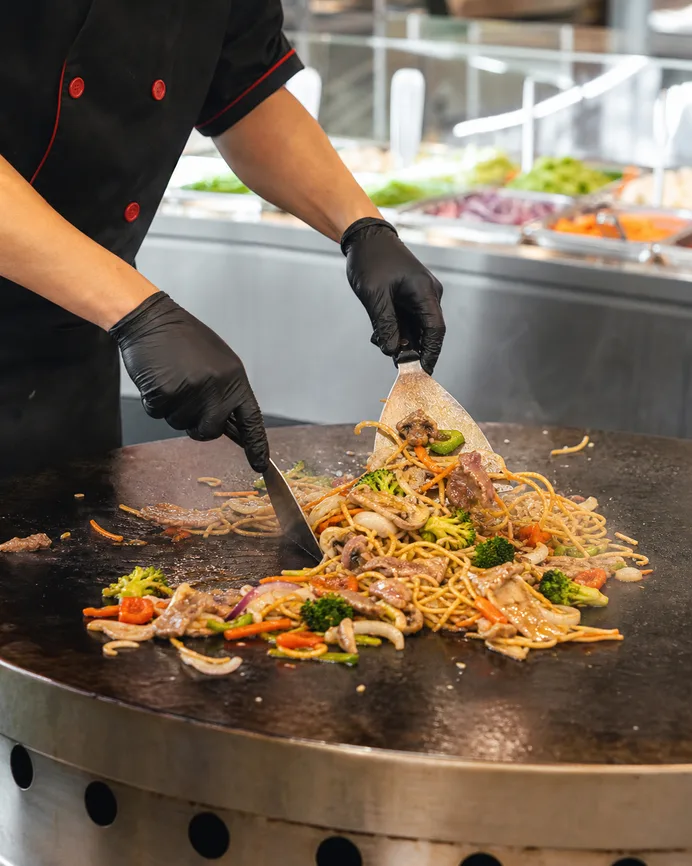 Chef cooking noodles, beef and vegetables on a round Mongolian grill